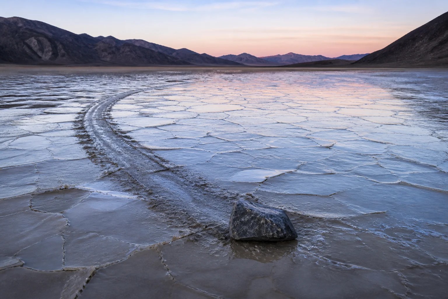 Editorial illustration of a dark rock, a shallow track, and thin ice on Racetrack Playa at dawn.
